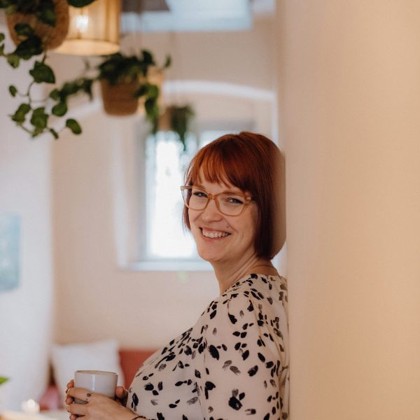 Karoline Krey, Coachin für persönliche Transformation bei marvelousme, lehnt mit einer Tasse in der Hand lächelnd an einer Wand in einem gemütlich eingerichteten Raum mit Hängepflanzen.