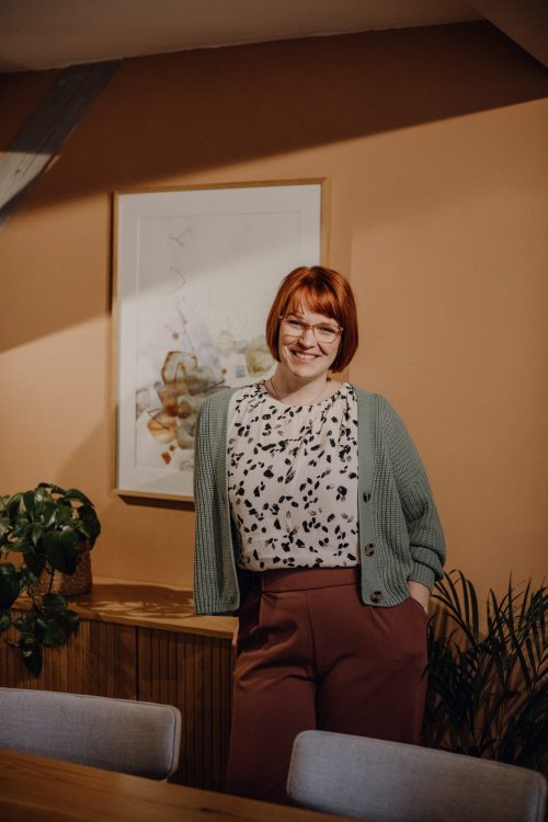 Karoline Krey, Coachin für persönliche Transformation bei marvelousme, steht lächelnd in einem warm gestalteten Coachingraum mit Pflanzen und Kunst an der Wand